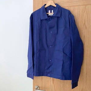 Könnte beinhalten: Eine einfarbige, königsblaue Arbeitsjacke mit klassischem Kragen, langen Ärmeln mit geknöpften Manschetten und einer Knopfleiste vorne. Die Jacke verfügt über eine Brusttasche und zwei aufgesetzte Taschen sowie ein weißes Etikett am Innenkragen.