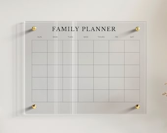 May include: A clear acrylic wall-mounted family planner calendar featuring a grid layout with seven columns labeled SUN through SAT. The header reads FAMILY PLANNER in black text. The board is secured to the wall with four gold-toned metal standoffs.