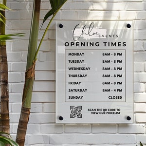 Op de afbeelding: Een wit bord met zwarte tekst met de openingstijden van Chloe Events. Het bord bevat een QR-code om de prijslijst te bekijken. Het bord is bevestigd aan een witte bakstenen muur.