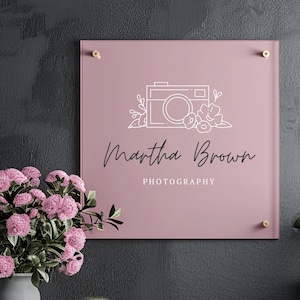 Könnte beinhalten: Ein quadratisches, rosa Schild mit dem Text "Martha Brown Photography" und einer Kamera-Grafik. Das Schild ist an einer dunkelgrauen Wand mit messingfarbener Hardware befestigt. Eine Vase mit rosa Blumen und ein kleiner Kaktus befinden sich im Vordergrund.