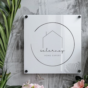 May include: White square sign with rounded corners and black text. The sign has a black circle with a house icon and the text "selernov" and "HOME EXPERT".