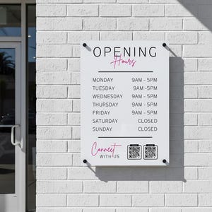 Peut inclure: Panneau rectangulaire blanc affichant les horaires d'ouverture d'un commerce, fixé sur un mur de briques blanches. Le panneau indique "OPENING Hours" en noir et rose, suivi d'une liste des jours et des heures. En dessous, "Connect with us" en rose, avec deux codes QR.