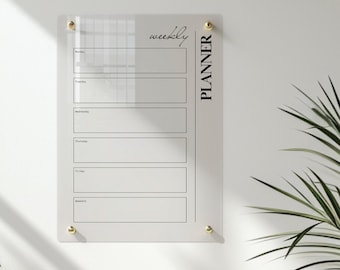 May include: A clear acrylic weekly planner board with gold-toned mounting hardware. The board features black printed text reading "weekly PLANNER" and six horizontal rectangular sections labeled Monday through Weekend for daily scheduling.