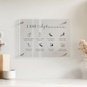 Peut inclure: Une plaque rectangulaire en acrylique transparent avec des fixations dorées, affichant le texte noir LASH Aftercare et huit icônes avec des instructions : éviter l'eau, les produits huileux, le mascara, le recourbe-cils, et conseils de sommeil.