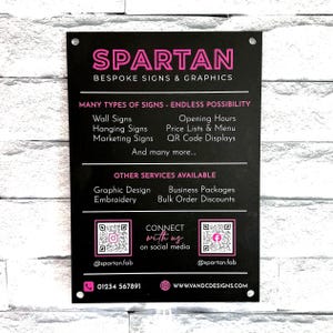 Peut inclure: Une enseigne rectangulaire noire avec du texte rose et blanc indiquant SPARTAN BESPOKE SIGNS & GRAPHICS. Elle liste des services comme les enseignes murales, les horaires et le design graphique, avec deux QR codes, réseaux sociaux et téléphone.