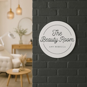 Peut inclure: Une pancarte ronde blanche avec du texte noir qui dit "The Beauty Room" avec le nom "Amy Rebecca" en dessous. La pancarte est montée sur un mur de briques noires.