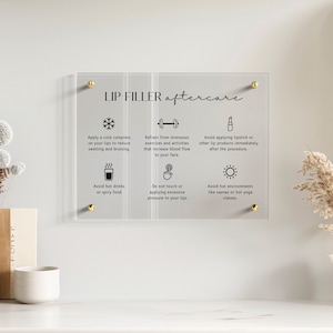 Peut inclure: Un panneau rectangulaire en acrylique transparent avec des fixations dorées, affichant le texte noir LIP FILLER aftercare et six icônes avec des instructions pour éviter les boissons chaudes, les exercices intenses, la pression et la chaleur.