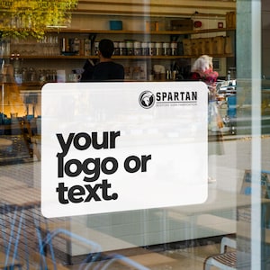 May include: A white rectangular sign with rounded corners is affixed to a glass window. The sign features the text "your logo or text" in bold, black font. Above the text is the "SPARTAN" logo and company name in black.
