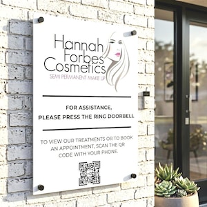 Könnte beinhalten: Ein rechteckiges weißes Schild für "Hannah Forbes Cosmetics" mit einer Grafik eines Frauengesichts. Es weist Besucher an, für Hilfe die Klingel zu betätigen und einen QR-Code für Termine zu scannen. Das Schild ist an einer weißen Backsteinmauer befestigt.