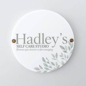 May include: A white, circular sign featuring the text "Hadley's SELF CARE STUDIO" in grey. The sign also includes the phrase "Because you deserve to feel amazing." Decorative green leaves and two brass-coloured rivets complete the design.