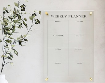 May include: A rectangular clear acrylic weekly planner board with black text and grid lines. The board features the heading WEEKLY PLANNER and sections for Monday through Sunday plus a Notes section. It is mounted with four gold-toned metal standoffs.