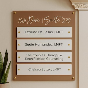 May include: A brown and white office directory sign with gold accents. The sign displays the address "1001 Dove | Suite 270" and lists four names and services: Czarina De Jesus, Sadie Hernández, The Couples Therapy & Reunification Counseling, and Chelsea Sutter.