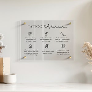 Peut inclure: Un panneau rectangulaire en acrylique dépoli avec des fixations dorées, affichant le texte noir TATTOO Aftercare et six icônes explicatives avec des instructions sur l'entretien des tatouages, le lavage, l'application de pommade et la protection solaire.