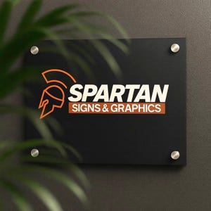 Könnte beinhalten: Ein schwarzes rechteckiges Schild mit dem Text "SPARTAN SIGNS & GRAPHICS" in Weiß und Orange. Das Schild zeigt ein orangefarbenes Spartanerhelm-Logo. Es ist mit vier silbernen Befestigungsbolzen an einer grauen Wand befestigt.