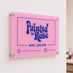 Könnte beinhalten: Ein rosa Acrylschild für ein Nagelstudio. Das Schild zeigt den Text "Painted by Rose" in einer dunkelblauen, stilisierten Schriftart mit den Worten "NAIL SALON" darunter. Das Schild enthält auch Social-Media-Handles und Website-Informationen.