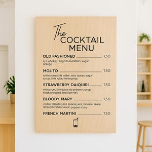 May include: A wooden cocktail menu sign with black text. The menu lists classic cocktails like Old Fashioned, Mojito, Strawberry Daiquiri, Bloody Mary, and French Martini. The sign is mounted on a white wall.