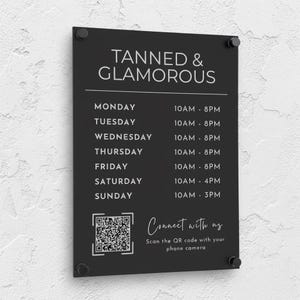 Peut inclure: Une enseigne professionnelle rectangulaire noire avec du texte blanc indiquant TANNED & GLAMOROUS, suivie d'un horaire hebdomadaire du lundi au dimanche, d'un code QR et du texte Connect with us. L'enseigne est fixée par quatre entretoises argentées.