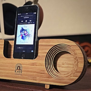 May include: A wooden phone stand with a circular cutout for sound amplification. The stand has a natural wood finish and the text "K.452 CRAFTS" is engraved on the front. A black smartphone is placed in the stand, displaying the music player with the song "Proef" by "Consciousuren" playing.