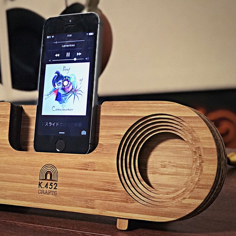 May include: A wooden phone stand with a circular cutout for sound amplification. The stand has a natural wood finish and the text "K.452 CRAFTS" is engraved on the front. A black smartphone is placed in the stand, displaying the music player with the song "Proef" by "Consciousuren" playing.