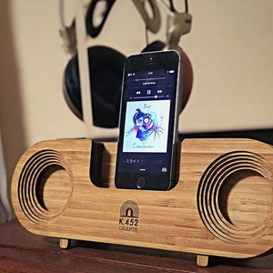 May include: A wooden speaker with a phone dock and a phone playing music. The speaker is made of bamboo and has a circular cutout for the speaker. The phone is a black iPhone with a screen showing the album art for "Proof of Consciousness" by "Roof". The speaker has the text "K.452 CRAFTS" on the front.