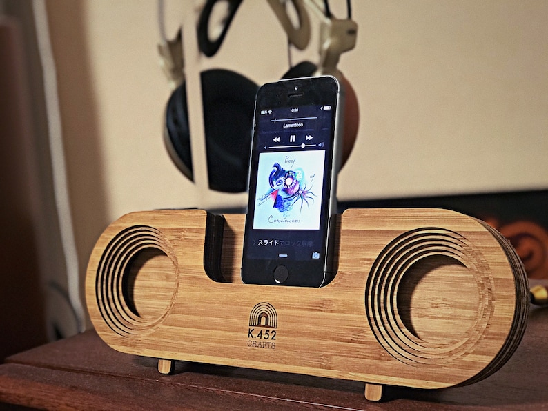May include: A wooden speaker with a phone dock and a phone playing music. The speaker is made of bamboo and has a circular cutout for the speaker. The phone is a black iPhone with a screen showing the album art for "Proof of Consciousness" by "Roof". The speaker has the text "K.452 CRAFTS" on the front.