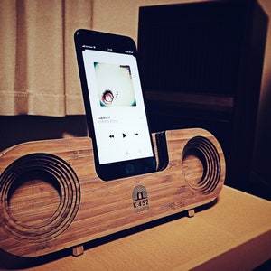 May include: A wooden speaker dock with a phone stand and two speakers. The dock is made of light brown wood and has a dark brown finish. The phone stand is made of black metal and has a slot for a phone. The speakers are made of black metal and have a circular shape. The dock has the text "K.452 CRAFTS" printed on the front.