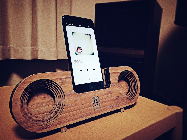 May include: A wooden speaker dock with a phone stand and two speakers. The dock is made of light brown wood and has a dark brown finish. The phone stand is made of black metal and has a slot for a phone. The speakers are made of black metal and have a circular shape. The dock has the text "K.452 CRAFTS" printed on the front.