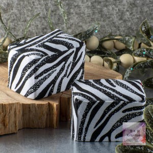 May include: Two square-shaped boxes with a black and white zebra print design. The boxes are made of fabric and are sitting on a wooden surface.