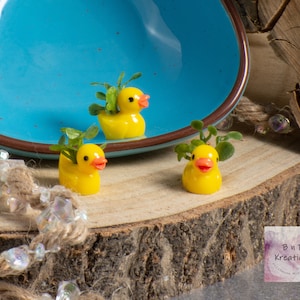 May include: Three miniature yellow rubber ducks with green plant accents. The ducks are sitting on a wooden surface with a blue bowl in the background.