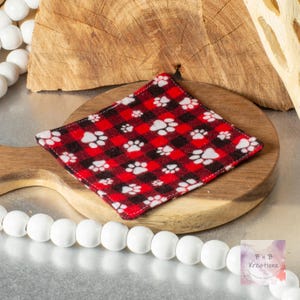 May include: A square, red and black plaid coaster with white paw prints. The coaster is made of fabric and is placed on a wooden cutting board. The background includes a wooden slice and white beads.