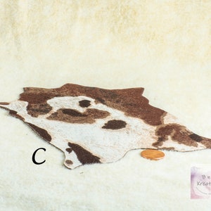 May include: A miniature rug with a cowhide pattern in shades of brown and white. The rug is irregularly shaped and has a textured appearance. A penny is placed next to the rug for size comparison. The letter "C" is printed on the rug.
