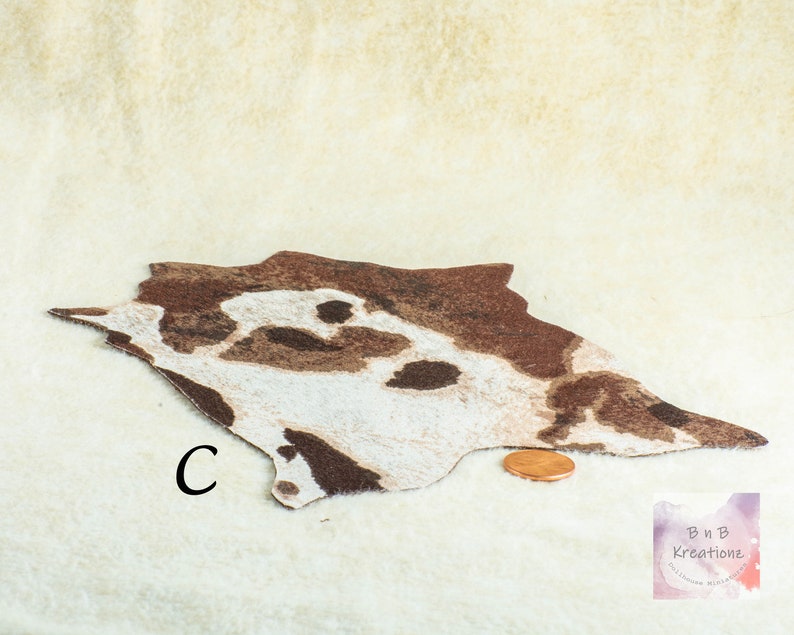 May include: A miniature rug with a cowhide pattern in shades of brown and white. The rug is irregularly shaped and has a textured appearance. A penny is placed next to the rug for size comparison. The letter "C" is printed on the rug.