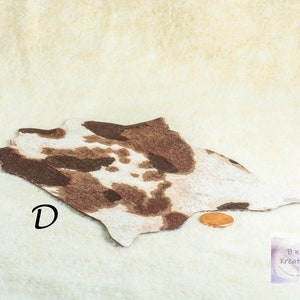 May include: A miniature cowhide rug with brown and white spots. The rug is irregularly shaped and lies on a cream-colored surface. A penny is next to the rug for scale. The letter "D" is printed on the rug.