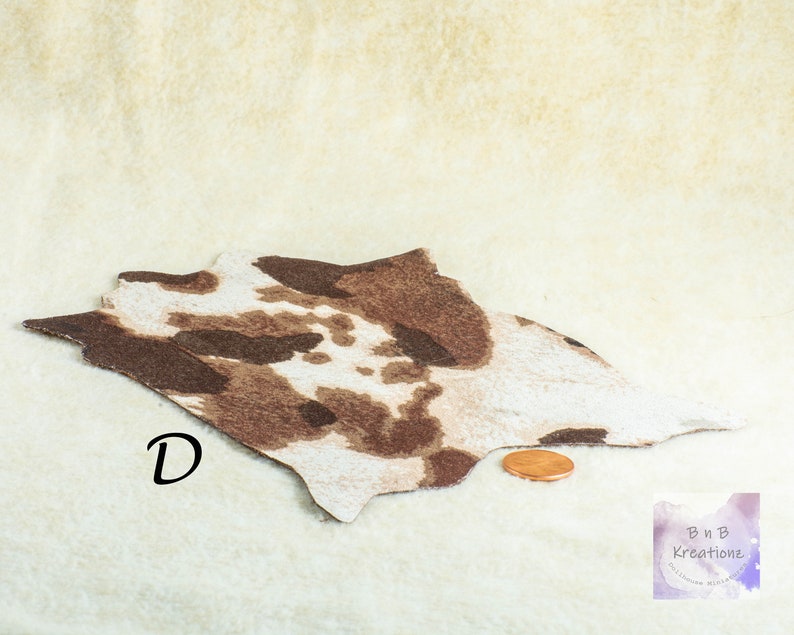 May include: A miniature cowhide rug with brown and white spots. The rug is irregularly shaped and lies on a cream-colored surface. A penny is next to the rug for scale. The letter "D" is printed on the rug.