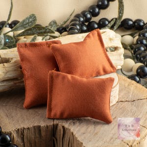 May include: Three small, rectangular, rust-colored fabric pillows. The pillows are arranged on a wooden surface, with a natural wood grain. The pillows are likely for jewelry or small item display. The pillows have a stitched border.