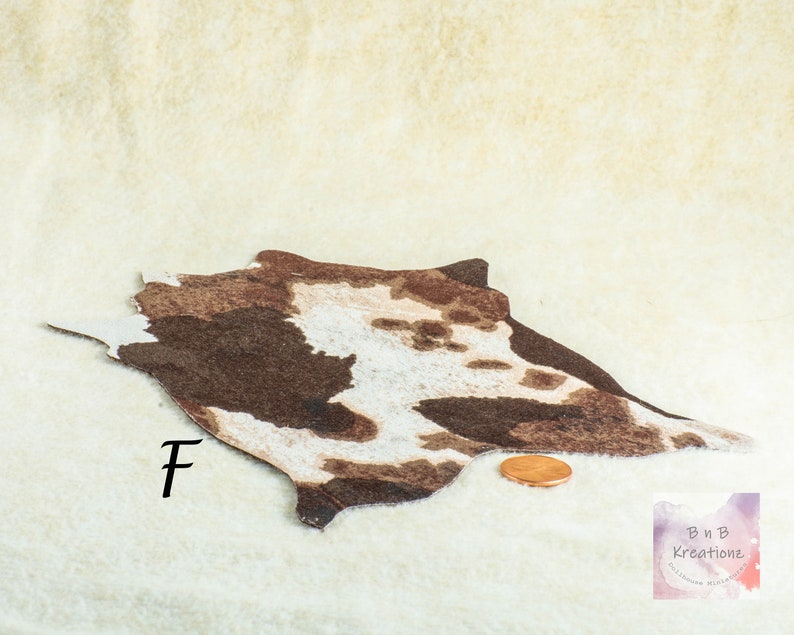 May include: A miniature cowhide rug with brown and white patterns. The rug is placed on a cream-colored surface. A penny is next to the rug for scale. The letter "F" is written on the rug.