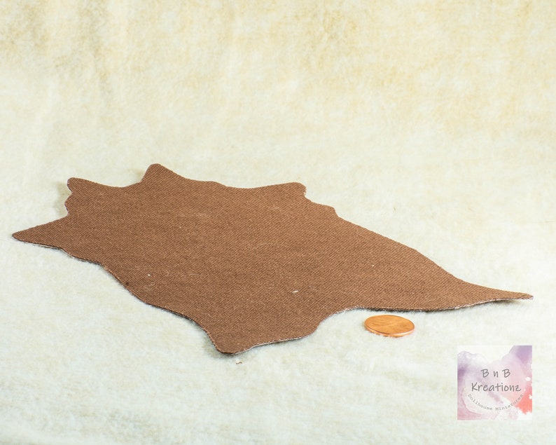 Shows the back side of a miniature cowskin rug. The fabric on the back is a brown canvas style texture. It is on a biege background. It has a penny in front of the rug to show scale.
They are made for 1:12 scale or 1:6 scale miniature dollhouses or diorama scenes and are not full sized.