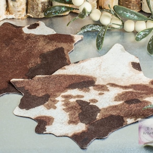 Shows a dollhouse miniature cowskin style rug in browns and cream. They are shown on a metal background with wooden beads. 
They are made for 1:12 scale or 1:6 scale miniature dollhouses or diorama scenes and are not full sized.

The logo for the shop that sells them, "B n' B Kreationz" is in the bottom righthand corner.