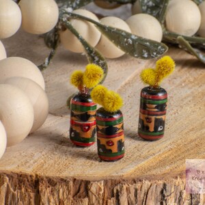 May include: Three small, hand-painted, cylindrical vases with black, green, red, and yellow designs. Each vase has a small, yellow, fuzzy flower on top.