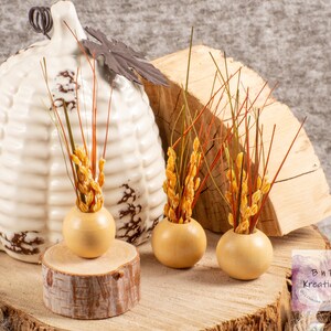 May include: Three small, round, wooden vases with a light brown finish. Each vase is filled with dried grasses and wheat stalks in shades of brown, orange, and yellow. The vases are arranged on a wooden slice.
