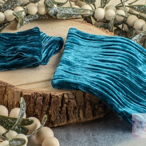 May include: Two pieces of teal blue velvet fabric with a crinkled texture. The fabric is folded and displayed on a wooden surface.