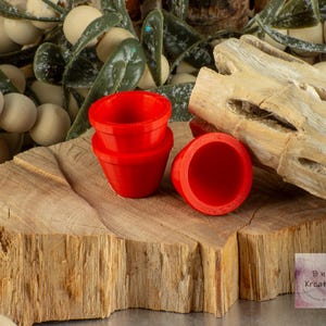 May include: Three red miniature flower pots stacked on top of each other on a wooden surface. The pots are made of a smooth, glossy material. The background is a blurred image of a branch with white berries.