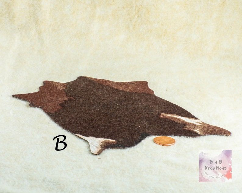 May include: Miniature dollhouse rug in a cowhide design. The rug is primarily brown with white and dark brown accents. A penny is next to the rug for scale. The letter "B" is printed on the rug. The rug is on a cream-colored surface.