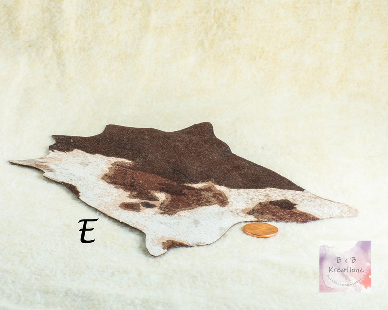 May include: A miniature cowhide rug with brown and white patterns. The rug is irregularly shaped and has a penny next to it for scale. The letter "E" is printed on the rug. The background is a cream-colored fabric.