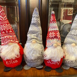 May include: Four Christmas gnomes with long, pointed hats and fluffy white beards. Two gnomes have red hats with white snowflake patterns, and two have gray hats. Each gnome has a name embroidered on its body: Elisa, Nanny, Joey, and Nicky.