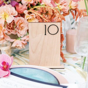 May include: A wooden table number with the number "10" in black lettering. The table number is sitting on a white table cloth with pink flowers in the background.