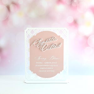 May include: A white laser-cut lattice frame holding a dusty rose sign with the text Signature Cocktail, Spring Bloom, and a list of ingredients including vodka, lemon juice, elderflower liqueur, simple syrup, and orange bitters.