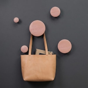 Op de afbeelding: Een beige leren tas met twee hengsels hangt aan een grote ronde houten wandhaak. In de tas zit een notitieboek met spiraalbinding. Vier extra ronde houten wandhaken in verschillende maten zijn bevestigd aan de donkergrijze muur.