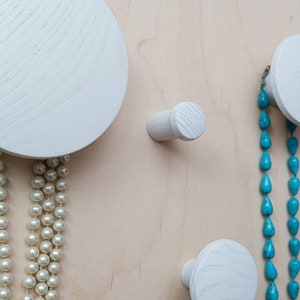 May include: White wooden wall hooks with a round shape and a cylindrical shape. A turquoise necklace hangs from one of the round hooks and a white pearl necklace hangs from the other round hook.
