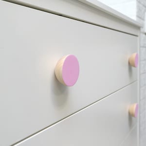 May include: Close-up of a white dresser with three drawers, each featuring a light pink and natural wood knob. The knobs are half-circle shaped and add a pop of color to the otherwise neutral furniture.